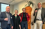 Fire personer står side om side innendørs og vender mot kamera. Tre av personene har yttertøy eller sekk. Bak dem står en stor, dekorativ gjenstand som ligner en gulrot med plante på toppen, og rommet har moderne belysning og lyse vegger.
