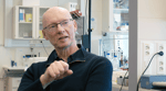 Middelaldrende mann uten hår med briller og marine genser sitter i et laboratorium og prater.
