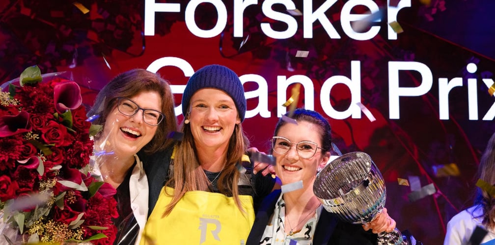 Tre personer står tett sammen på en scene omgitt av konfetti. Den ene holder en stor blomsterbukett, mens en annen holder et sølvfarget trofé. I bakgrunnen vises deler av teksten «Forsker Grand Prix».