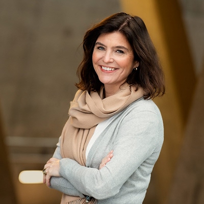 Kjersti Løken Stavrum med armene i kors, iført lys grå cardigan og beige skjerf. Står foran en innendørs bakgrunn med uklare arkitektoniske elementer.