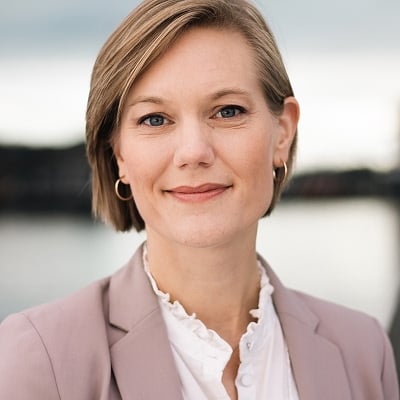 Bilde av Sigird Bratlie. Hun har på seg en lys lilla blazer og hvit bluse og står foran vann og uskarpe strukturer i bakgrunnen.