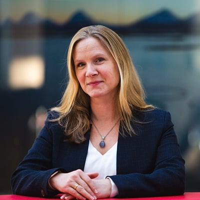 Bilde av Mari Sundli Tveit. Hun har  langt hår og mørk blazer og sitter ved et bord i et kontormiljø.