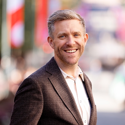 Geir Christian står utendørs iført en mørkebrun blazer og hvit skjorte. I bakgrunnen skimtes et urbant miljø med fargerike bannere og mennesker i det fjerne. Personen smiler og ser rett mot kameraet.