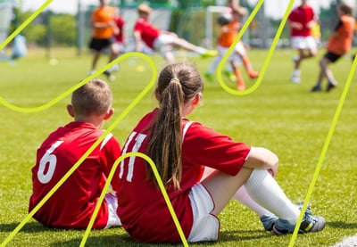 To barn i røde fotballdrakter med numrene 6 og 11 sitter på gresset ved sidelinjen og ser på en fotballkamp eller trening. I bakgrunnen spiller flere barn fotball på en grønn bane.