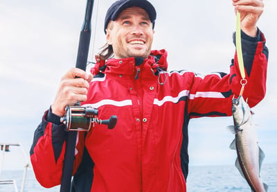 Person i rød jakke holder en fisk i snøre og en fiskestang om bord på en båt på havet.