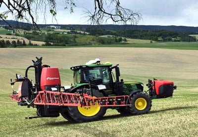 Traktor med åkersprøyte arbeider på jordet i et åpent kulturlandskap med åkre og skog i bakgrunnen.