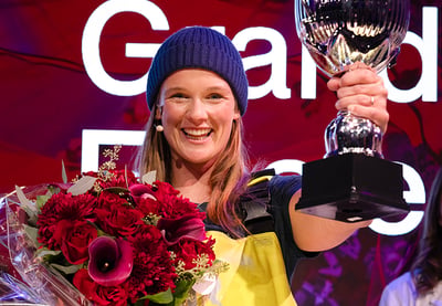 Person på scene holder opp en pokal og en stor bukett røde blomster, foran en bakgrunn med teksten «Grand Prix»