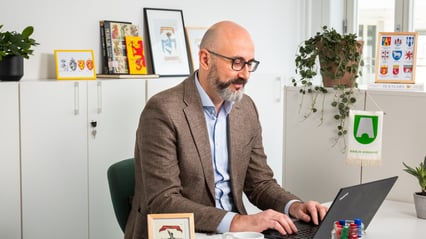 En mann med skjegg sitter i et kontorlandskap foran en PC.