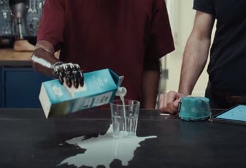 En hånd heller melk fra en kartong ned i et tomt glass på et bord, men mye av melken renner utenfor og søles på bordflaten. Hånden som holder kartongen er mørk og skinnende, det er en robotarmprotese. 