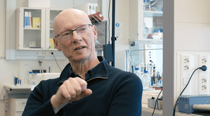 Middelaldrende mann uten hår med briller og marine genser sitter i et laboratorium og prater.