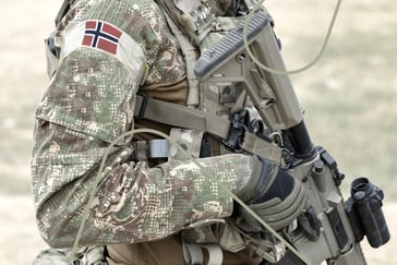 Bildet viser utsnitt av en soldat med det norske falgget på skulderen mens han holder et automatvåpen.