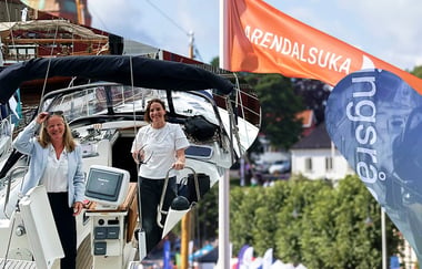 To personer står om bord i en seilbåt ved kai under Arendalsuka. I bakgrunnen ses bygninger og arrangementstelt. Til høyre i bildet er et oransje flagg med teksten «ARENDALSUKA».