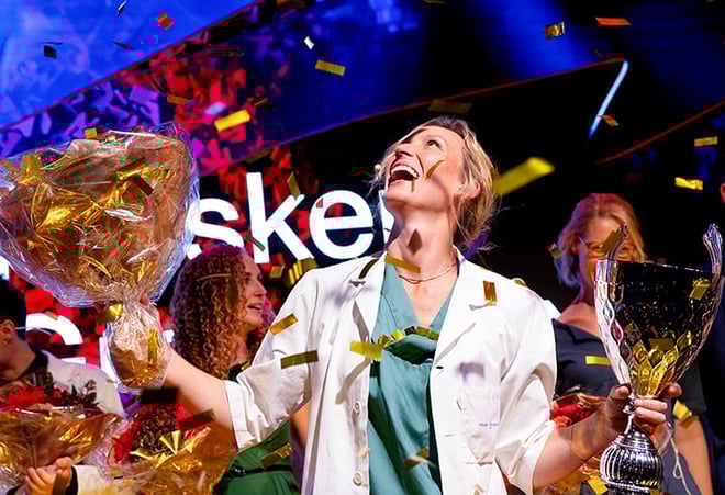 En person i hvit frakk og grønne klær holder en stor blomsterbukett og et trofé. Rundt personen faller det konfetti, og andre personer med blomsterbuketter er delvis synlige i bakgrunnen.