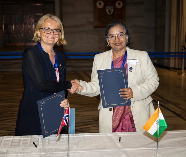 To personer håndhilser over et bord mens de holder hvert sitt signerte dokument. På bordet står små flagg fra Norge og India.