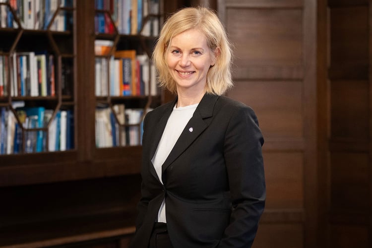 Foto av kvinne med halvlangt, lyst hår, i svart dressjakke og hvit skjorte, står foran en bokhylle fylt med bøker. Hun smiler til kameraet.