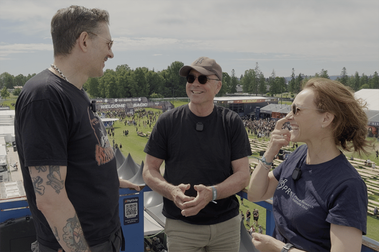 to menn og en kvinne prater sammen, festivalområde i bakgrunnen