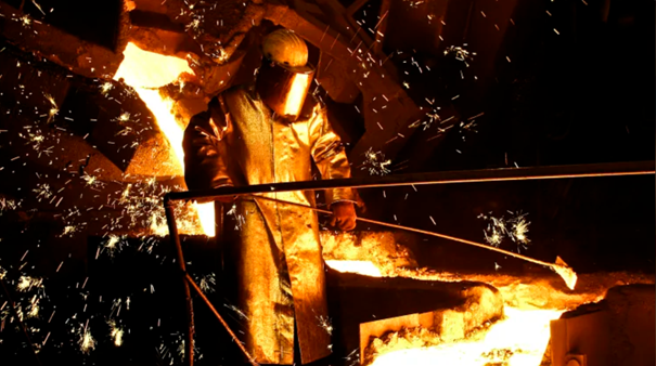 Person med vernedrakt og vernehjelp jobber med flytende metaller i et veldig varmt rom. Det flytende metallet lyser opp personen.