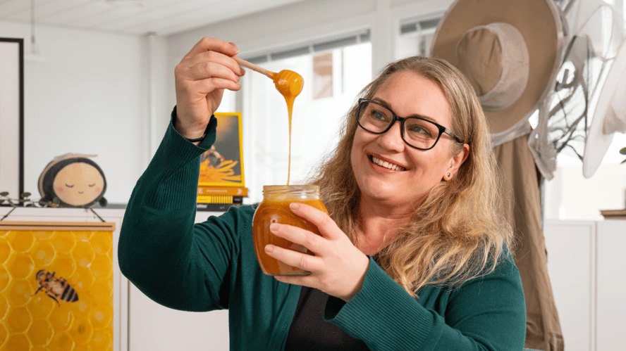 Blid dame med briller og lyst, langt hår holder opp honning fra et glass