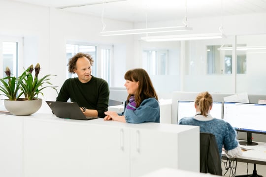 Tre ansatte på kontor. To samtaler rundt en bærbar PC, mens en annen jobber foran to PC-skjermer.