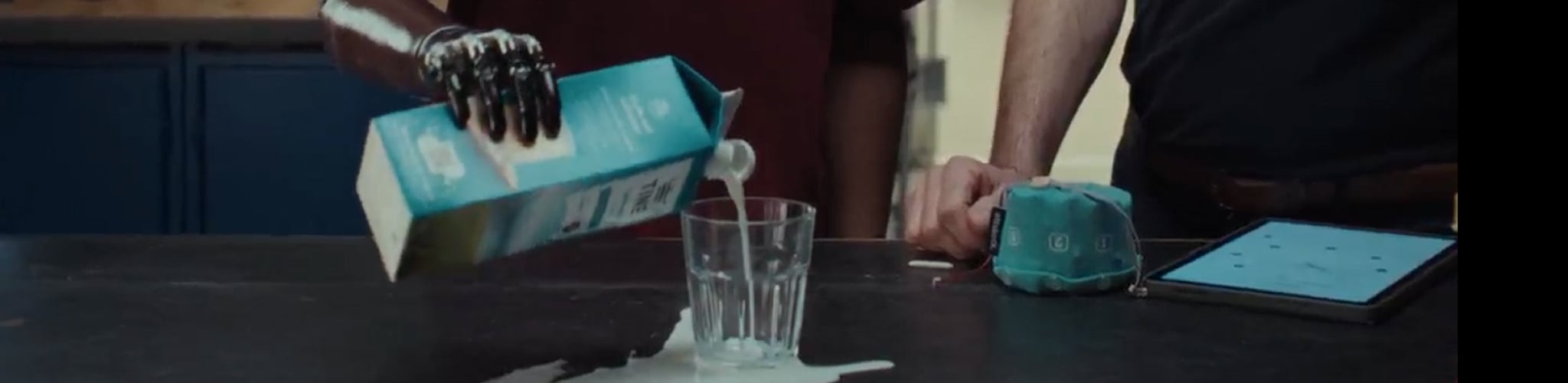 En h&aring;nd heller melk fra en kartong ned i et tomt glass p&aring; et bord, men mye av melken renner utenfor og s&oslash;les p&aring; bordflaten. H&aring;nden som holder kartongen er m&oslash;rk og skinnende, det er en robotarmprotese. 