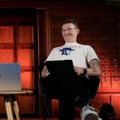 En mann sitter i en stol på en scene med mørk bakgrunn. Han ser lurt i kamera mens han smiler. Ved siden av seg har han et bord med en bærbar PC.