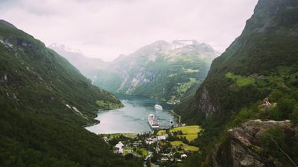 Fjord med grønne fjellsider og sne på fjelltoppene. Innerst i fjorden er det en bygd der det ligger det et cruise skip til kai