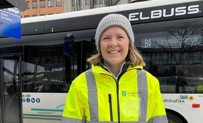 Smilende kvinne med refleksgul jakke står foran elbuss.