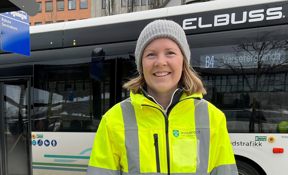 Smilende kvinne med refleksgul jakke står foran elbuss.